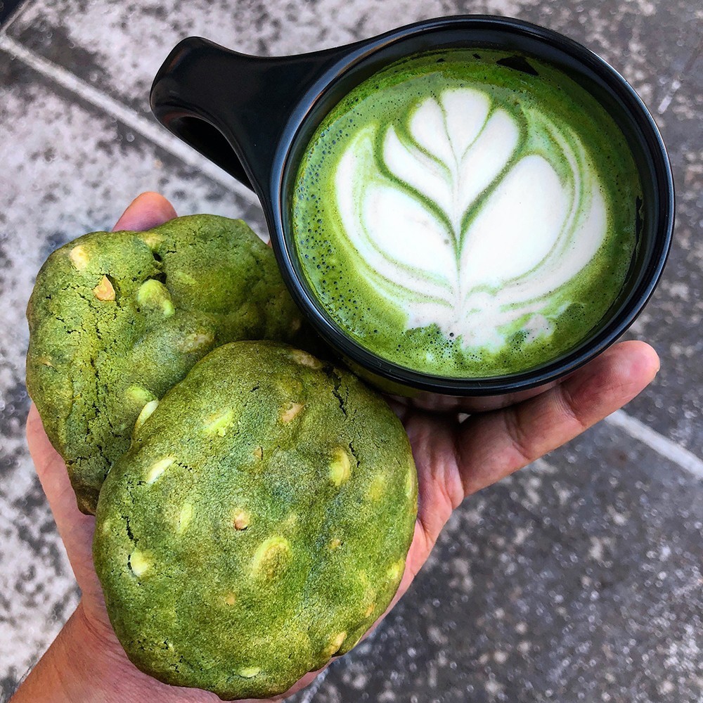 Dear Mama offers coffee, espresso, and specialty drinks, as well as an assortment of fresh-baked pastries and other light fare. A hand holding two large matcha cookies and a matcha latte