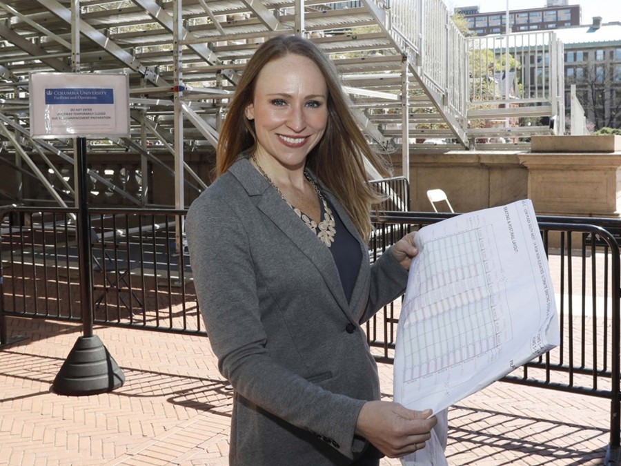 Mary Waldorf smiling next to the Commencement bleachers, holding documents detailing the setup
