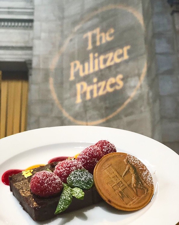A brownie and garnish on a plate next to a wall with an illuminated logo that says The Pulitzer Prizes