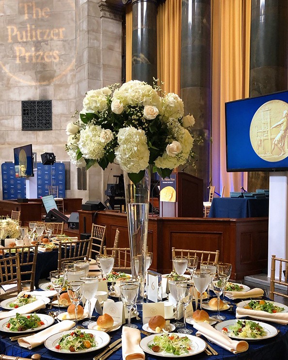 A table set up with plates, flowers, food, with the Pulitzer Prize logos on blue screens in the background