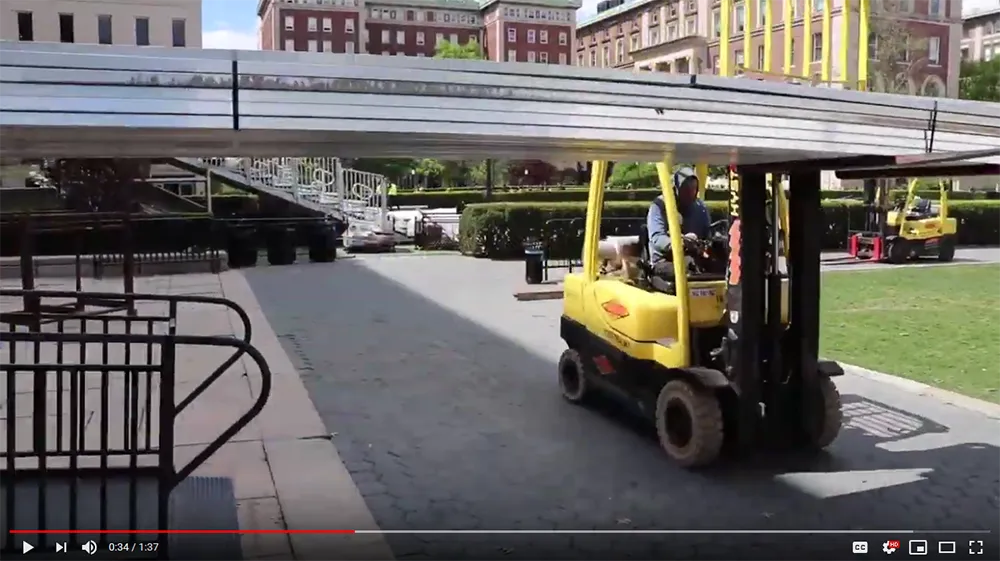 A screenshot of the video Building Commencement, showing a forklift carrying bleachers