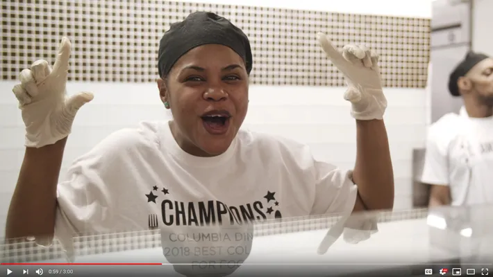A still of a video on Columbia Dining winning the Daily Meal award, with a Columbia Dining employee exciting and holding up #1 sign with her hands