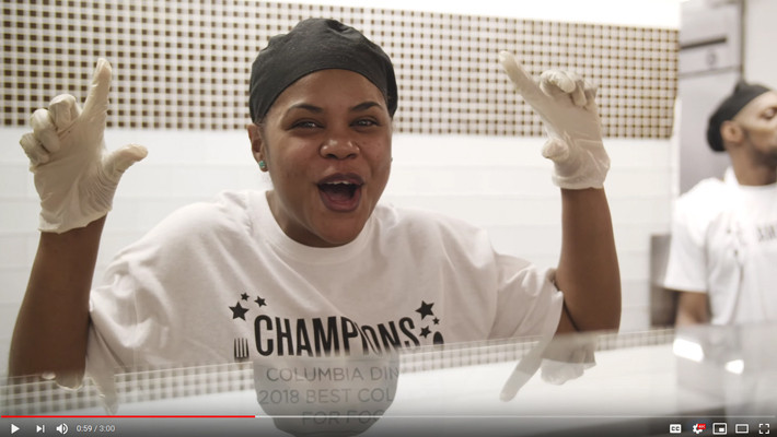 A still of a video on Columbia Dining winning the Daily Meal award, with a Columbia Dining employee exciting and holding up #1 sign with her hands
