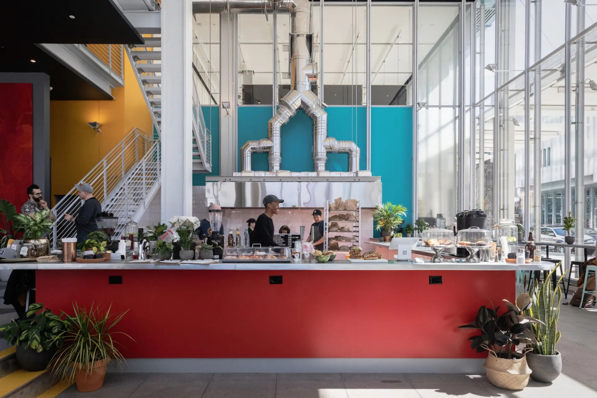 Dear Mama Coffee's cafe in the Jerome L. Greene Science Center with a red counter with plants on the ground and an assortment of pastries on top, with teal and yellow walls in the background and a white staircase.