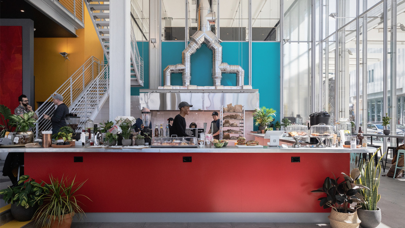 Dear Mama Coffee's cafe in the Jerome L. Greene Science Center with a red counter with plants on the ground and an assortment of pastries on top, with teal and yellow walls in the background and a white staircase.